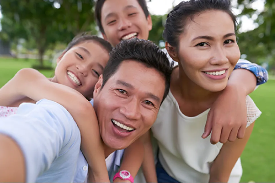 Happy Asian family smiling outdoors taking a close-up selfie together in a green park on a sunny day.