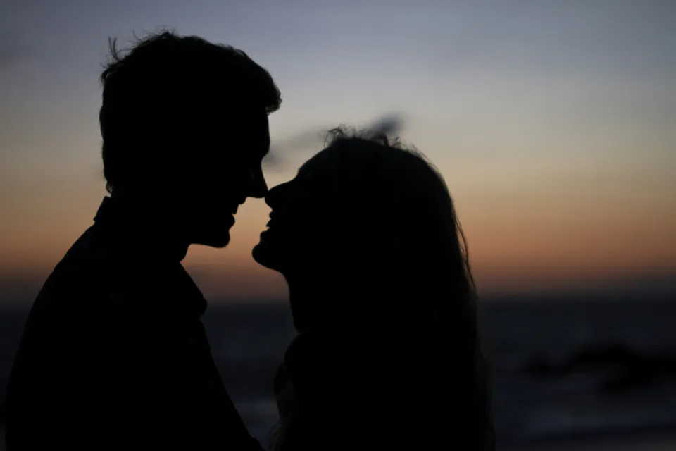 Silhouette of a couple facing each other at sunset by the ocean, sharing a romantic moment.