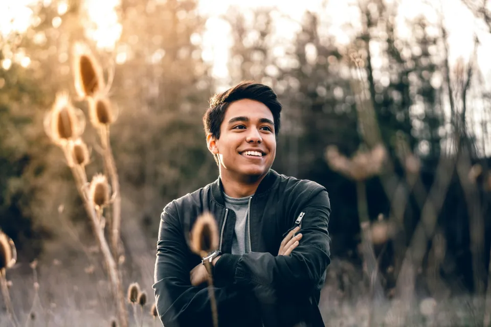 Smiling young man in black jacket standing outdoors with arms crossed during golden hour in nature.