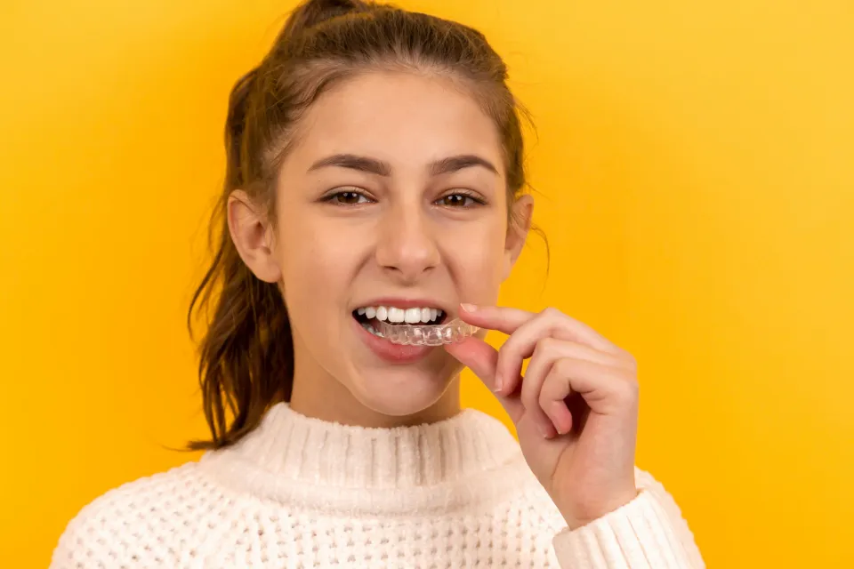 Young woman in white sweater placing a clear dental aligner on her teeth against yellow background