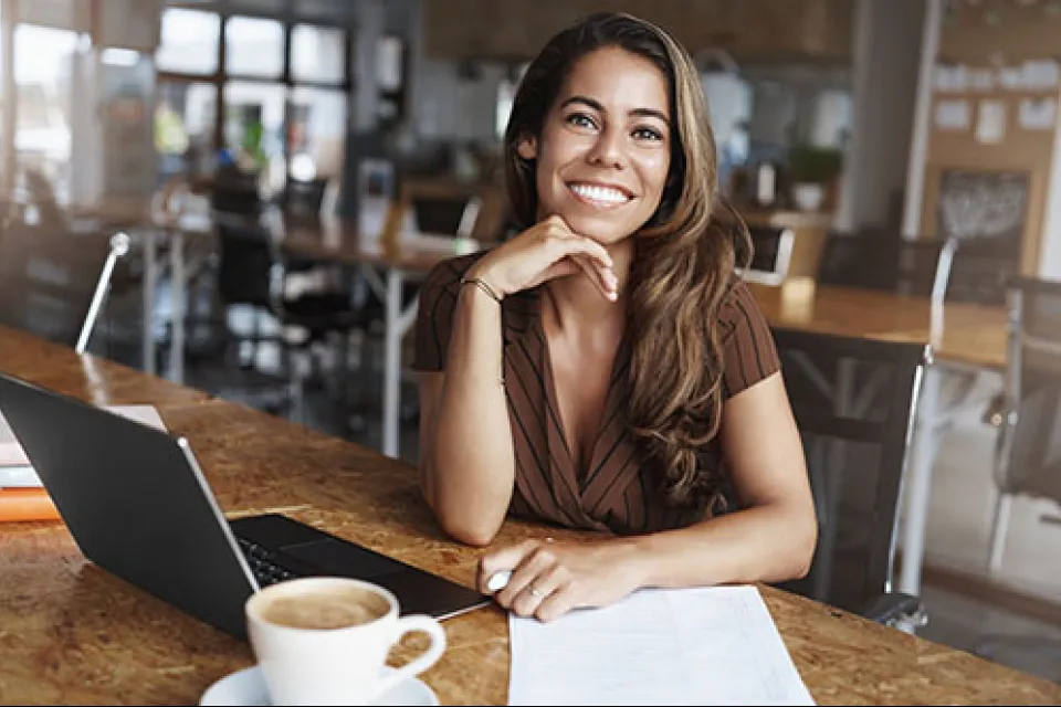 Smiling woman sitting at wooden desk with laptop, coffee cup, and papers in modern office space.