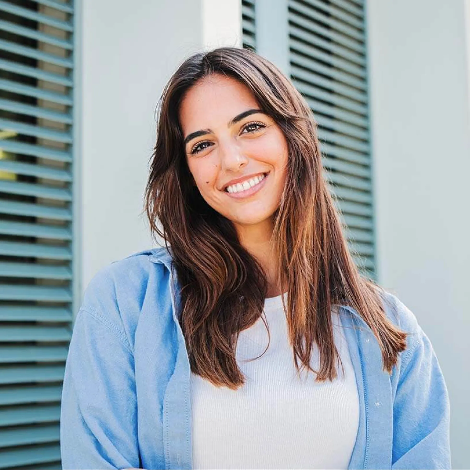 Smiling woman with long hair stands in front of a modern building with green shutters.