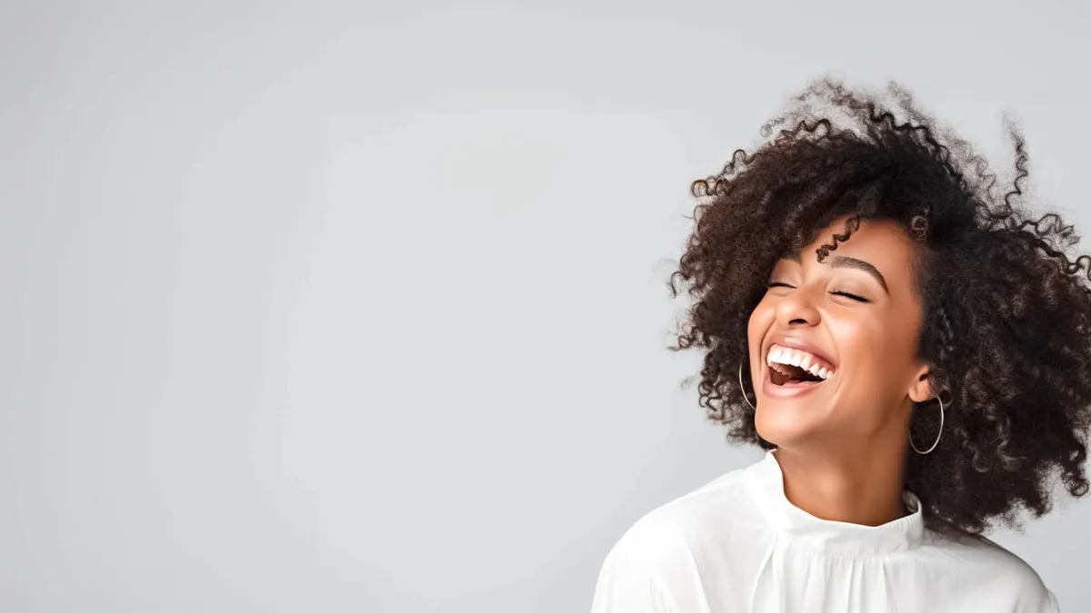 Happy woman with curly hair smiling against a gray background, radiating joy and positivity.