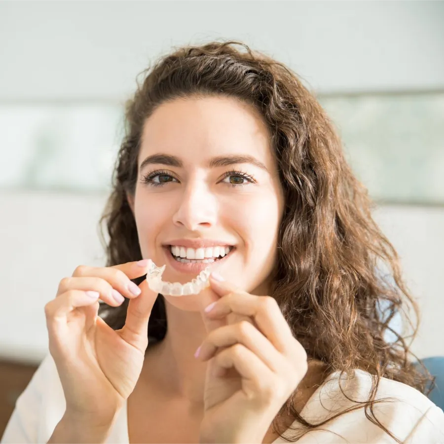 Young smiling woman holding a clear dental aligner near her mouth in a bright room.