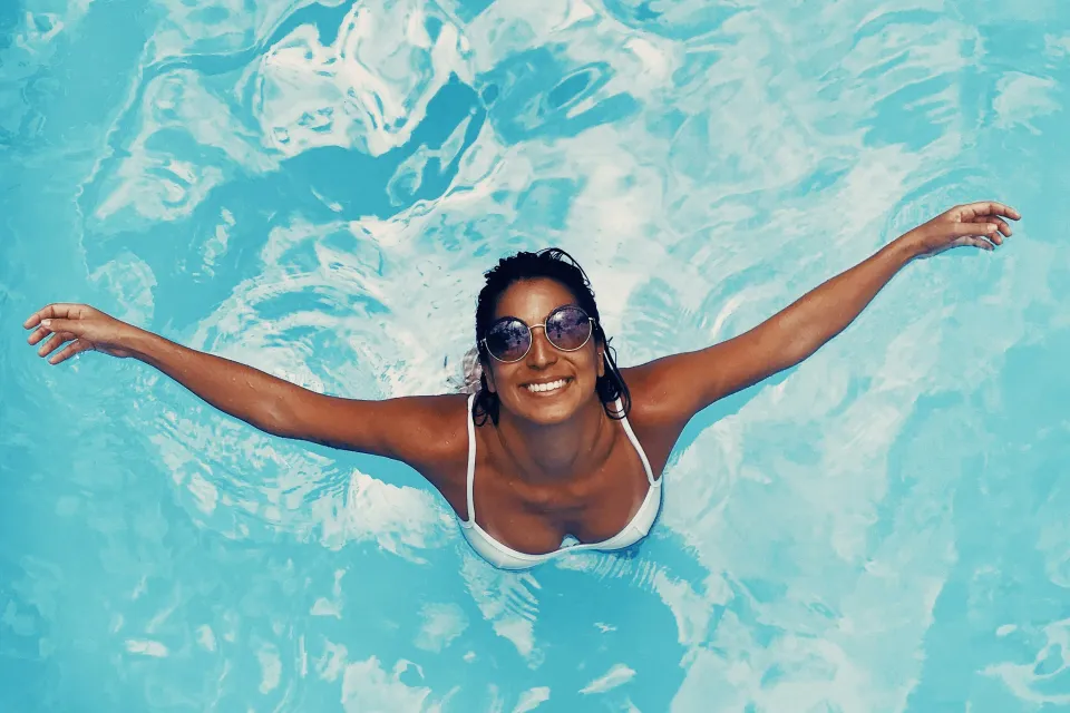 Smiling woman wearing sunglasses and white bikini top floating with arms outstretched in clear blue pool water.