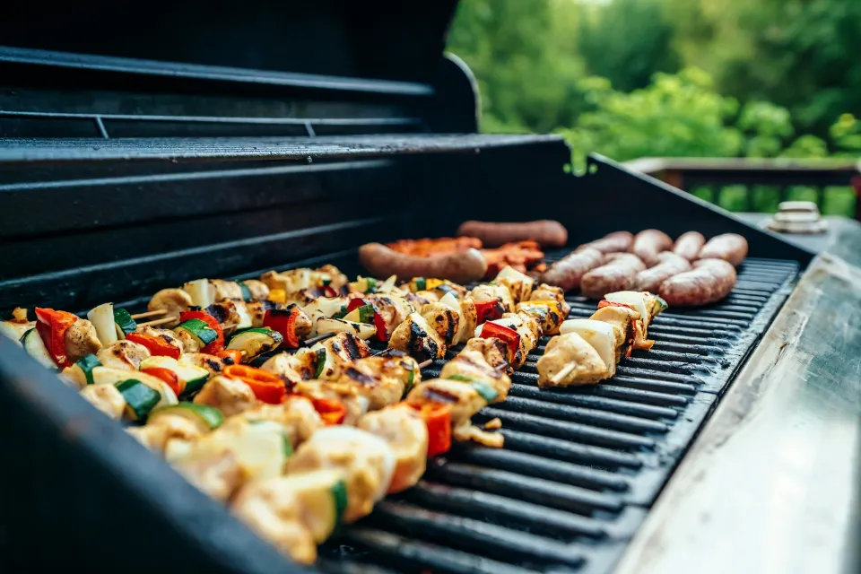 Grilled chicken and vegetable skewers with sausages cooking on an outdoor barbecue grill during daytime.