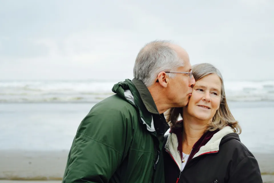 Older man in green jacket kisses smiling woman on cheek at a cloudy beach with waves in the background.