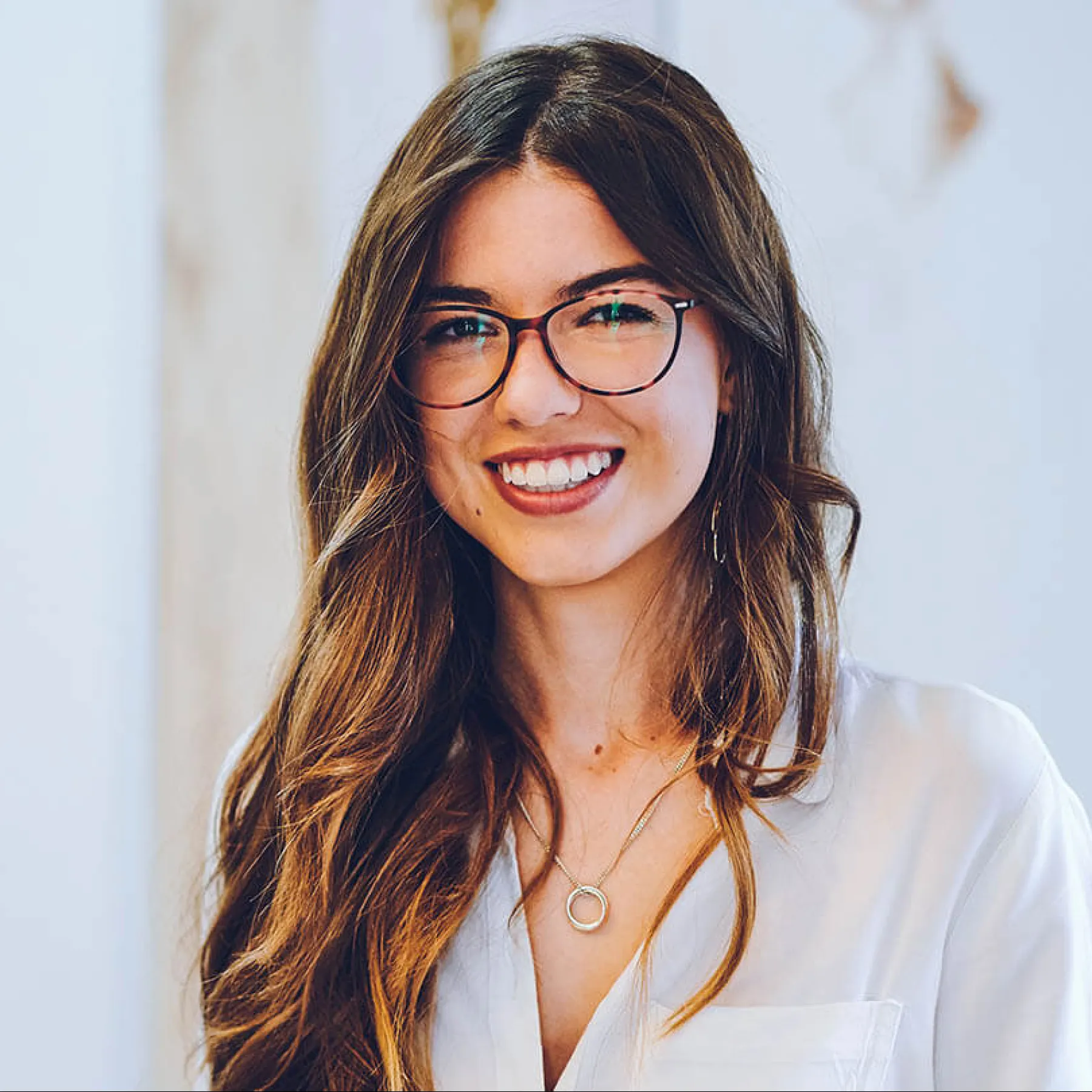 Smiling woman with long hair and glasses, wearing a white blouse in a bright indoor setting.