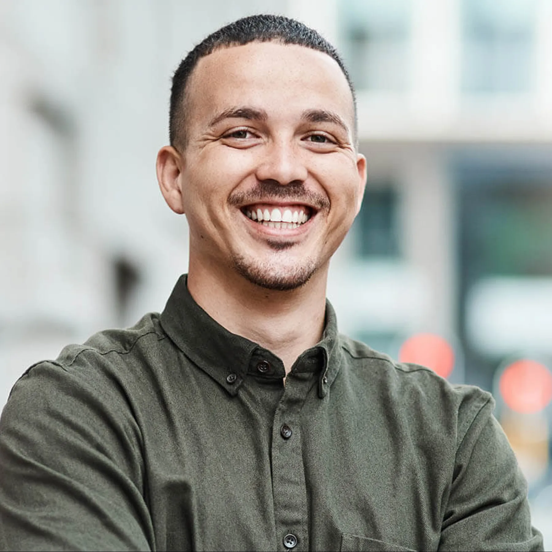 Smiling man in green shirt stands confidently outdoors with blurred city background.