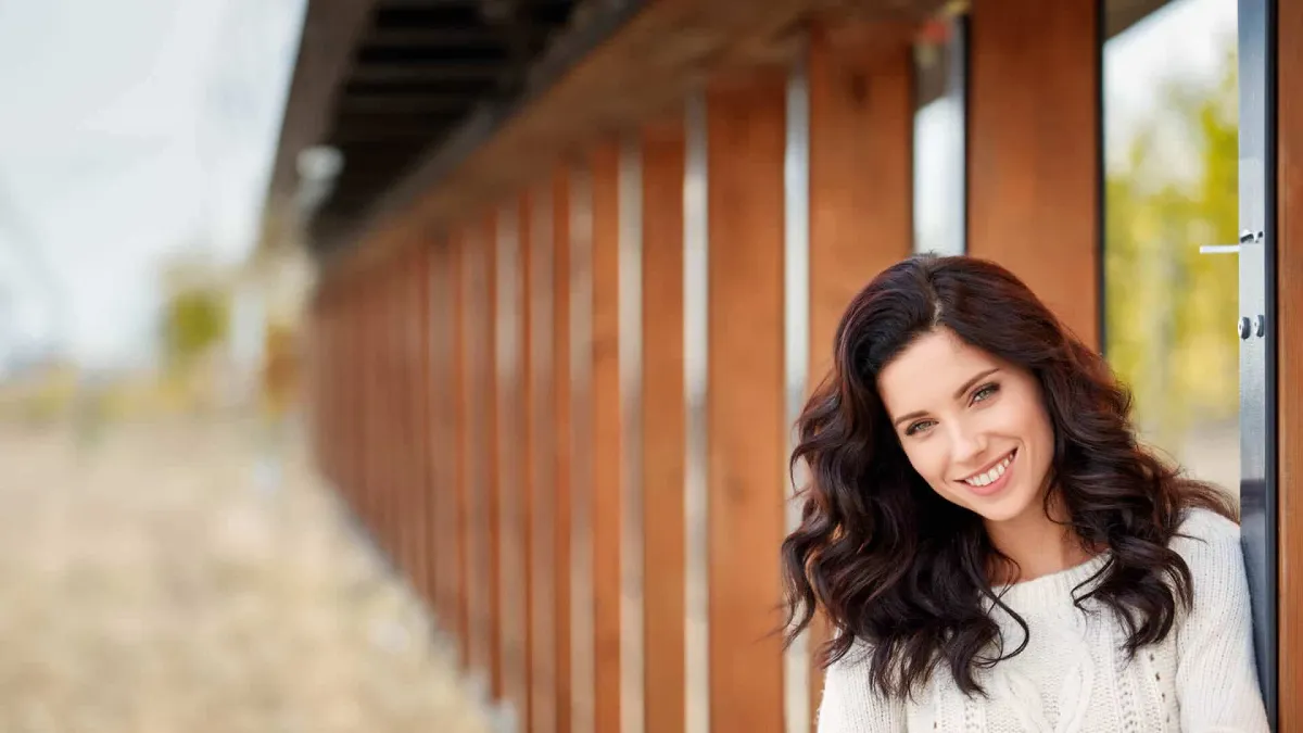 Smiling woman in a cozy white sweater near wooden structure in a serene outdoor setting.